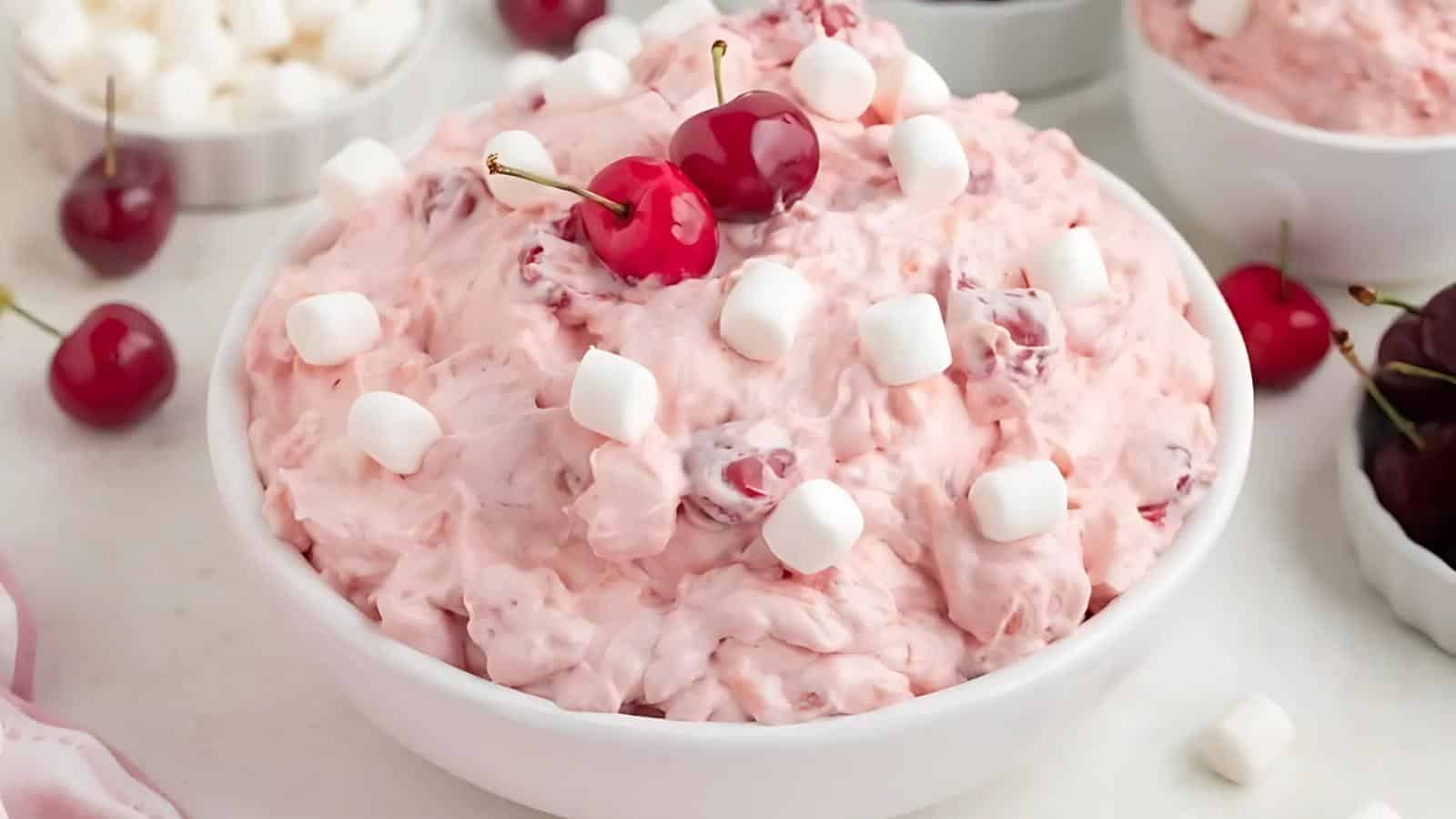 A white bowl filled with pink cherry fluff salad topped with mini marshmallows and cherries, with additional bowls of cherries and marshmallows in the background.