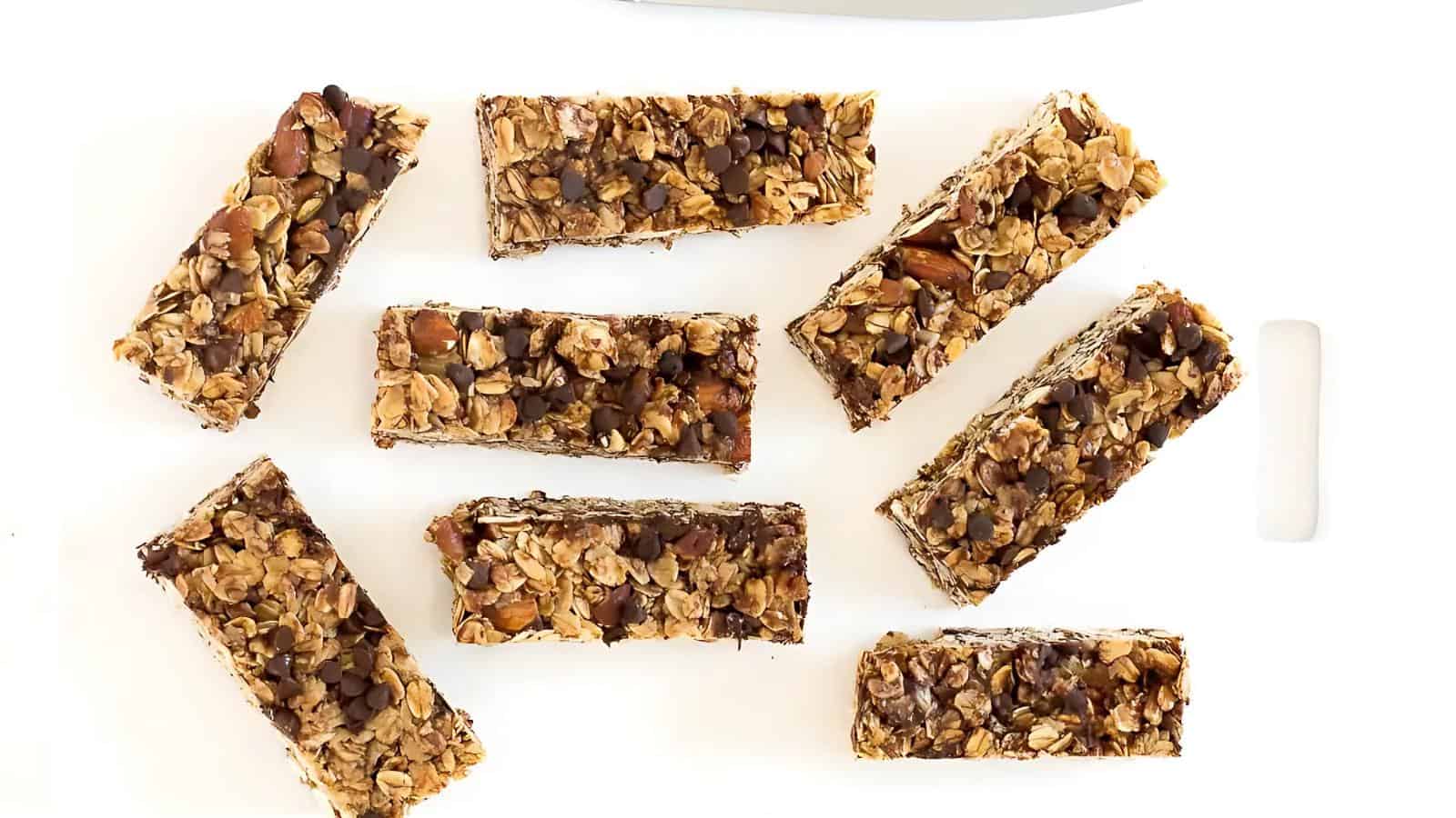 Eight homemade granola bars with oats, nuts, and chocolate chips are arranged on a white cutting board.