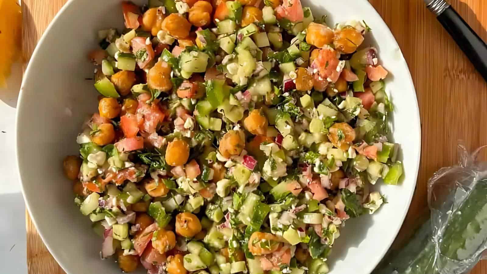 A white bowl filled with a chopped salad containing cucumbers, tomatoes, chickpeas, green peppers, and herbs, placed on a wooden surface.
