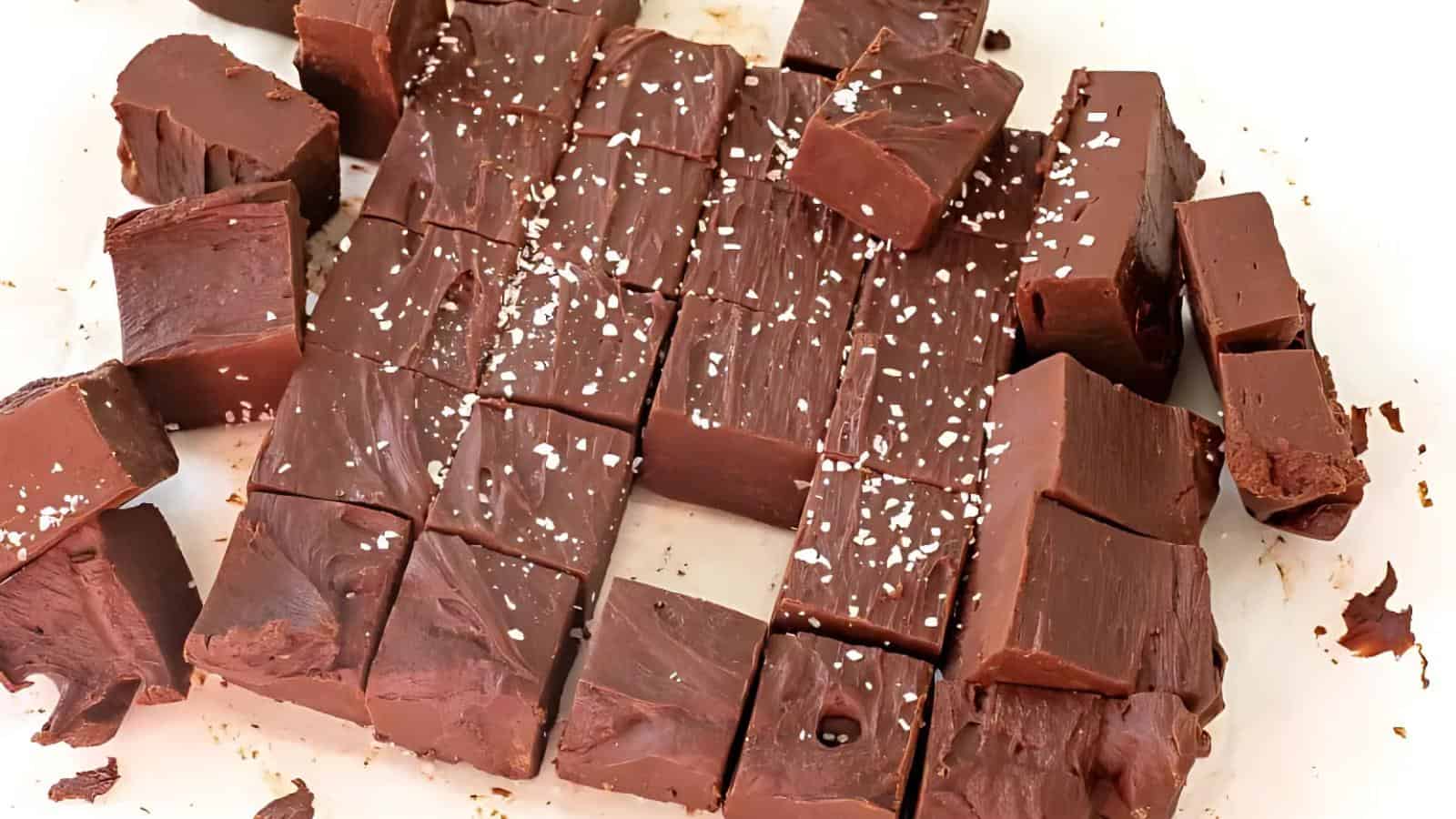 Squares of chocolate fudge topped with coarse sea salt are arranged on a white surface.