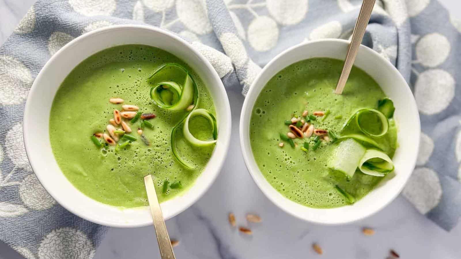 Two bowls of creamy green soup topped with pine nuts and cucumber ribbons, each with a spoon, placed on a patterned cloth.