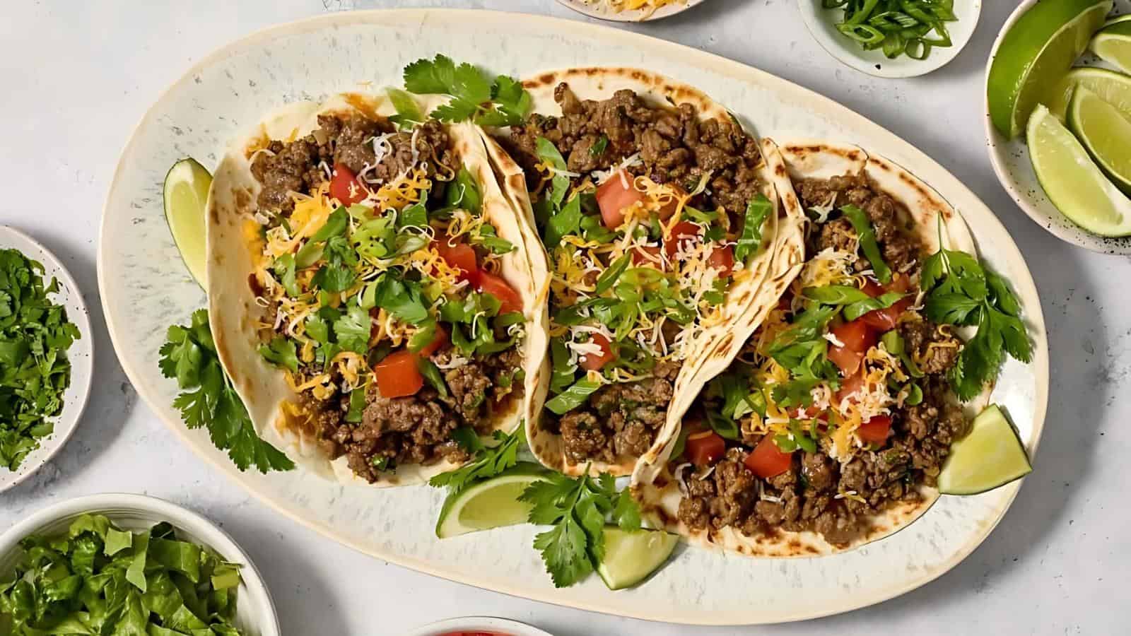 A platter with three beef tacos topped with shredded cheese, chopped tomatoes, and cilantro, served with lime wedges and surrounded by bowls of fresh toppings.