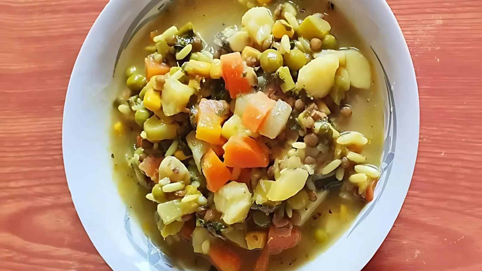 A bowl of vegetable soup containing diced carrots, potatoes, peas, and other assorted vegetables in a clear broth, on a wooden table.