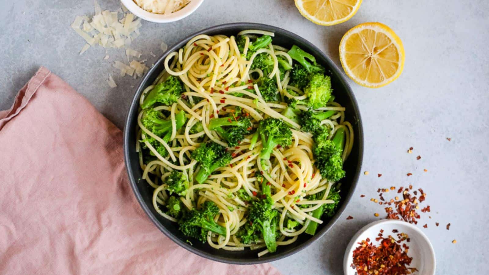 A bowl of spaghetti with broccoli, garnished with red pepper flakes, next to lemon halves, grated cheese, and a pink napkin on a gray surface.