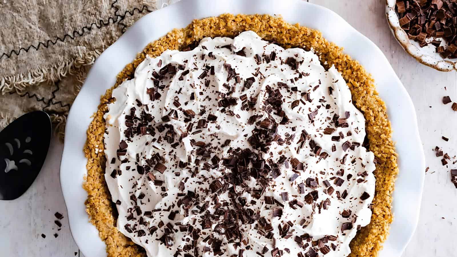 A cream pie with a crumb crust, topped with whipped cream and chocolate shavings, served on a white plate.