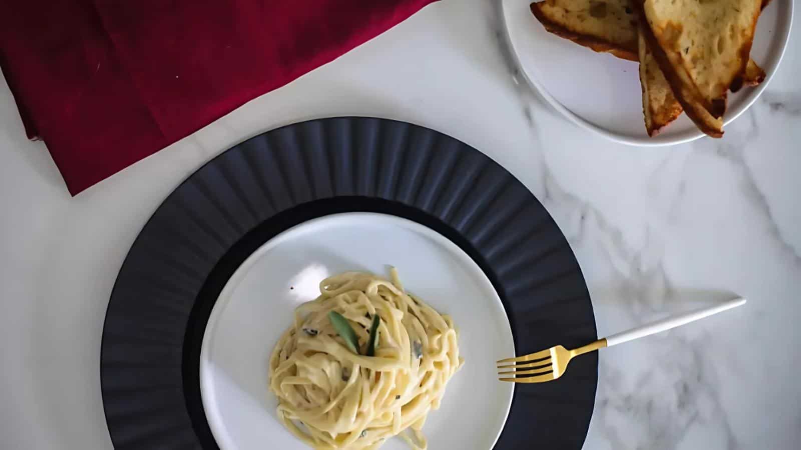 A plate of creamy pasta with herbs and a gold-handled fork sits on a black charger, next to a plate of toasted bread on a marble surface with a red cloth.