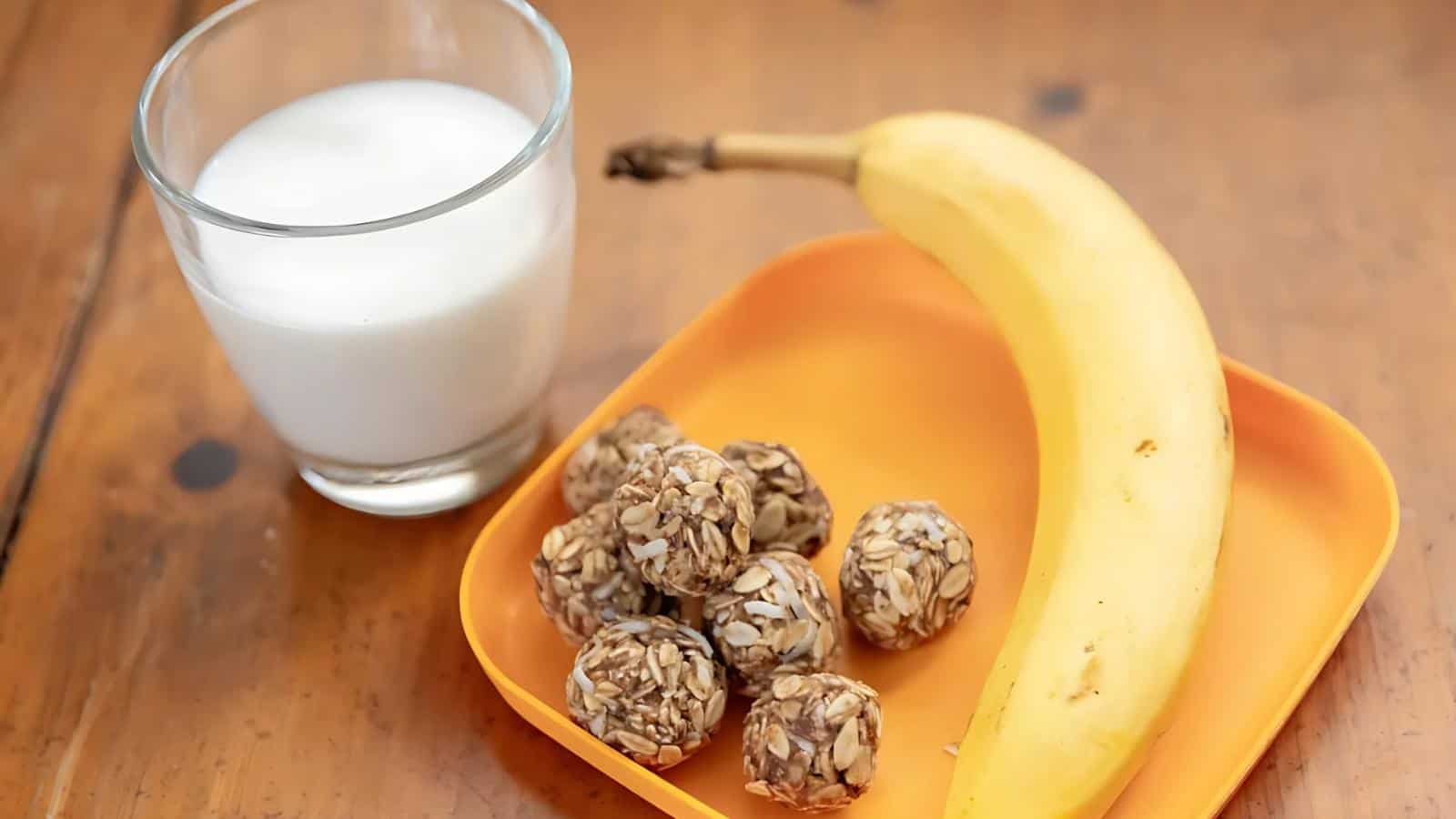 A glass of milk, a whole banana, and several oat and seed snack balls are arranged on an orange plate on a wooden surface.