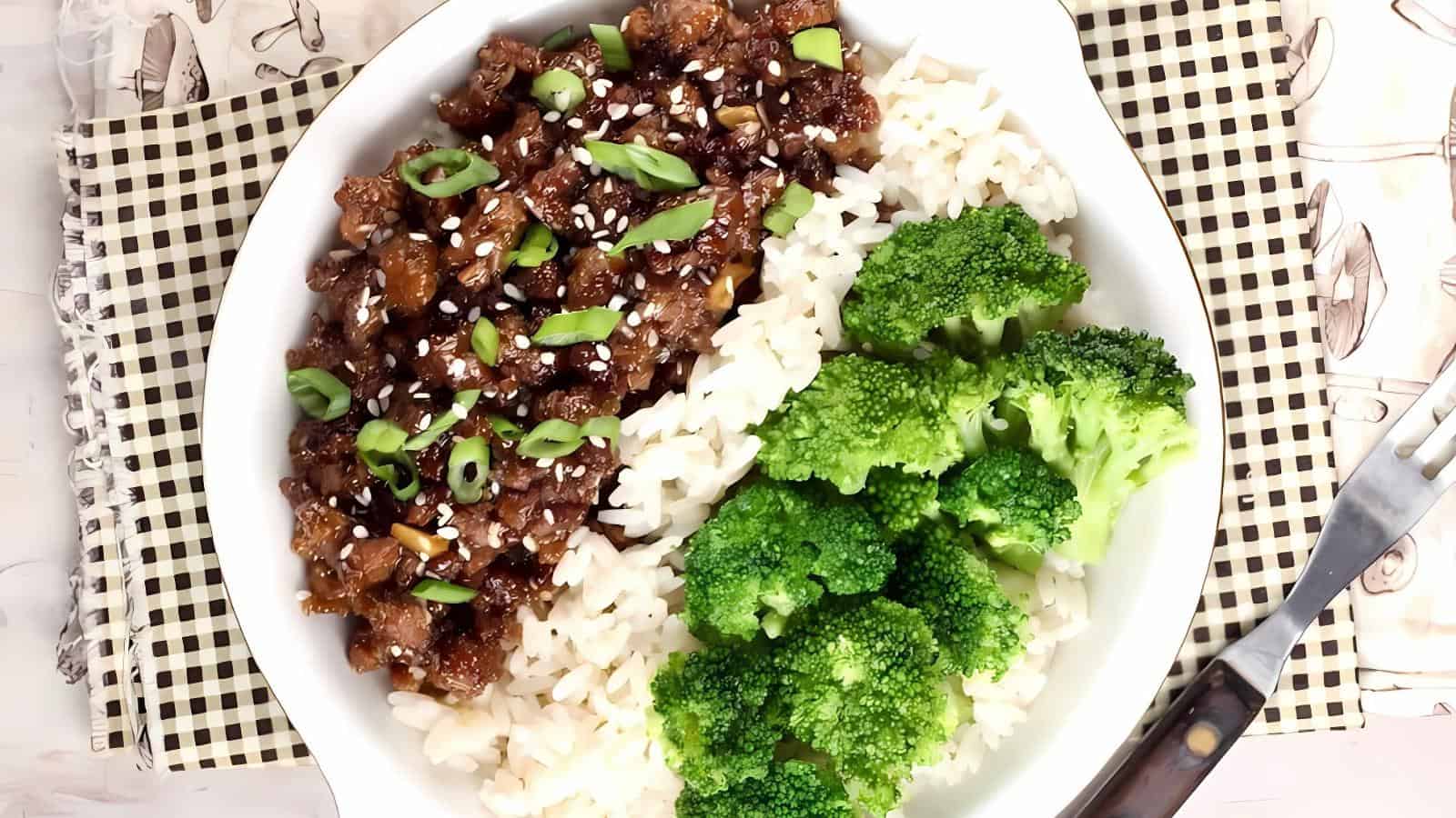 A white bowl with white rice, cooked broccoli florets, and a serving of saucy beef topped with sesame seeds and chopped green onions.