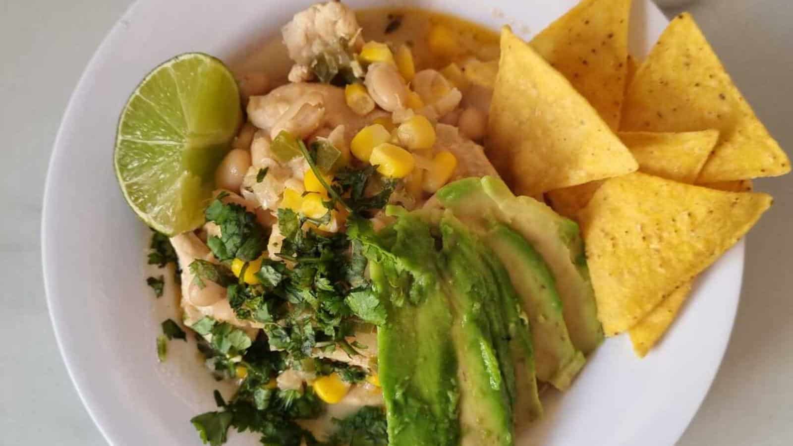 A bowl of white chili topped with sliced avocado, chopped cilantro, corn, and a lime wedge, served with tortilla chips on the side.