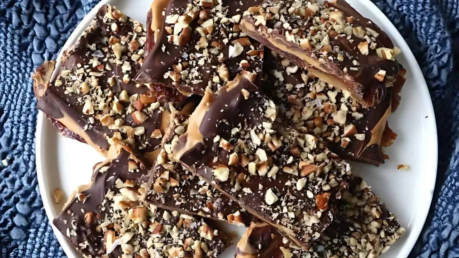 Pieces of chocolate toffee bark topped with chopped nuts are piled on a white plate.