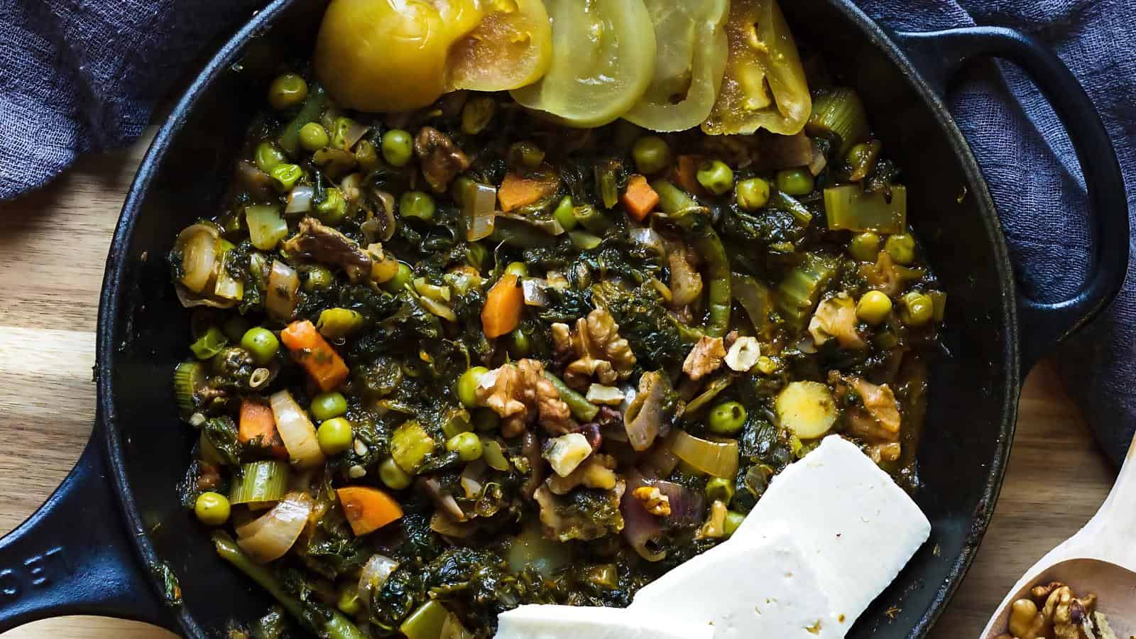 A skillet filled with a vegetable and walnut stew, garnished with yellow tomatoes and served with slices of white cheese.