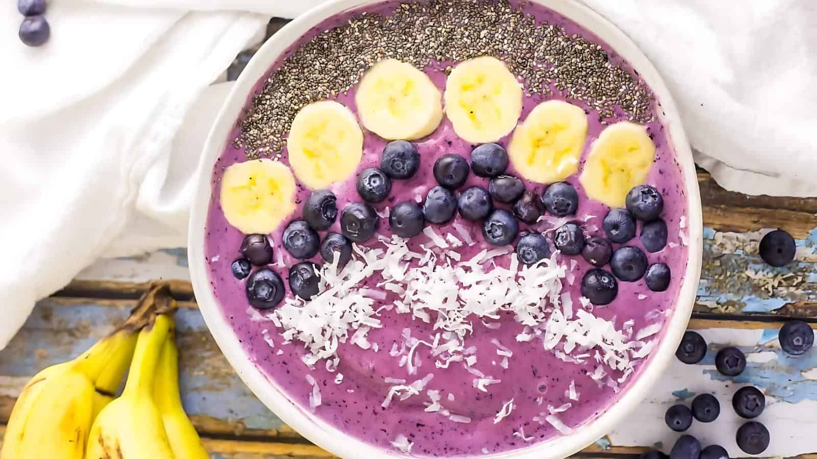 A smoothie bowl topped with chia seeds, banana slices, blueberries, and shredded coconut, placed on a wooden surface beside a bunch of bananas.