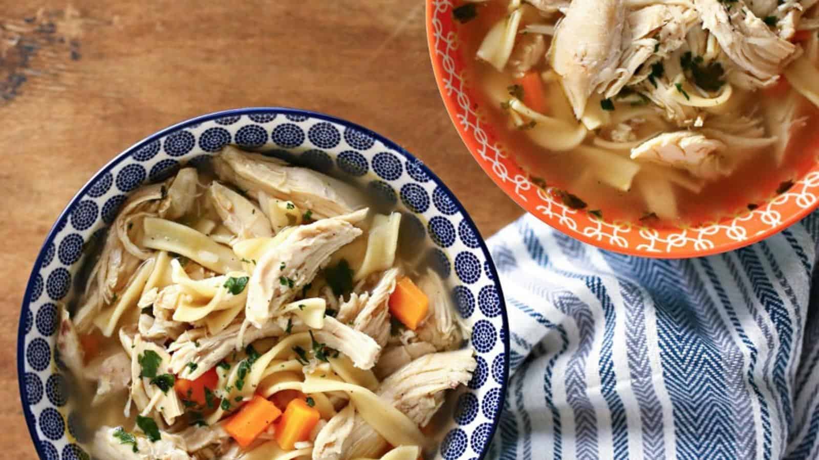 Two bowls of chicken noodle soup with shredded chicken, carrots, noodles, and herbs on a wooden surface beside a blue-and-white striped cloth.