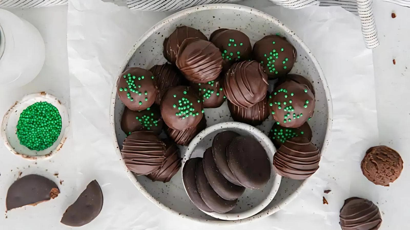 A plate of chocolate-covered cookies, some decorated with green sprinkles, sits on a white surface with more cookies and a bowl of green sprinkles nearby.