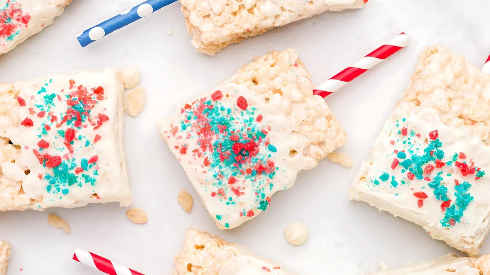 Rice Krispies treats coated with white chocolate, topped with red and blue sprinkles, each on a striped straw, arranged on a white surface.