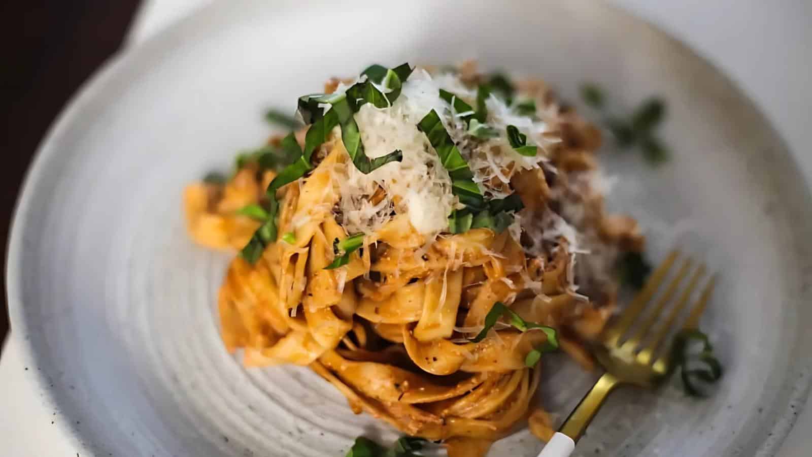 A plate of fettuccine pasta with tomato sauce, topped with grated cheese and chopped basil, with a gold fork beside the serving.