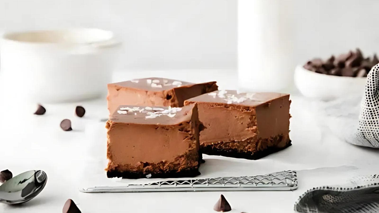 Three chocolate cheesecake bars with a chocolate crust are arranged on a cooling rack, with chocolate chips scattered nearby and blurred bowls in the background.