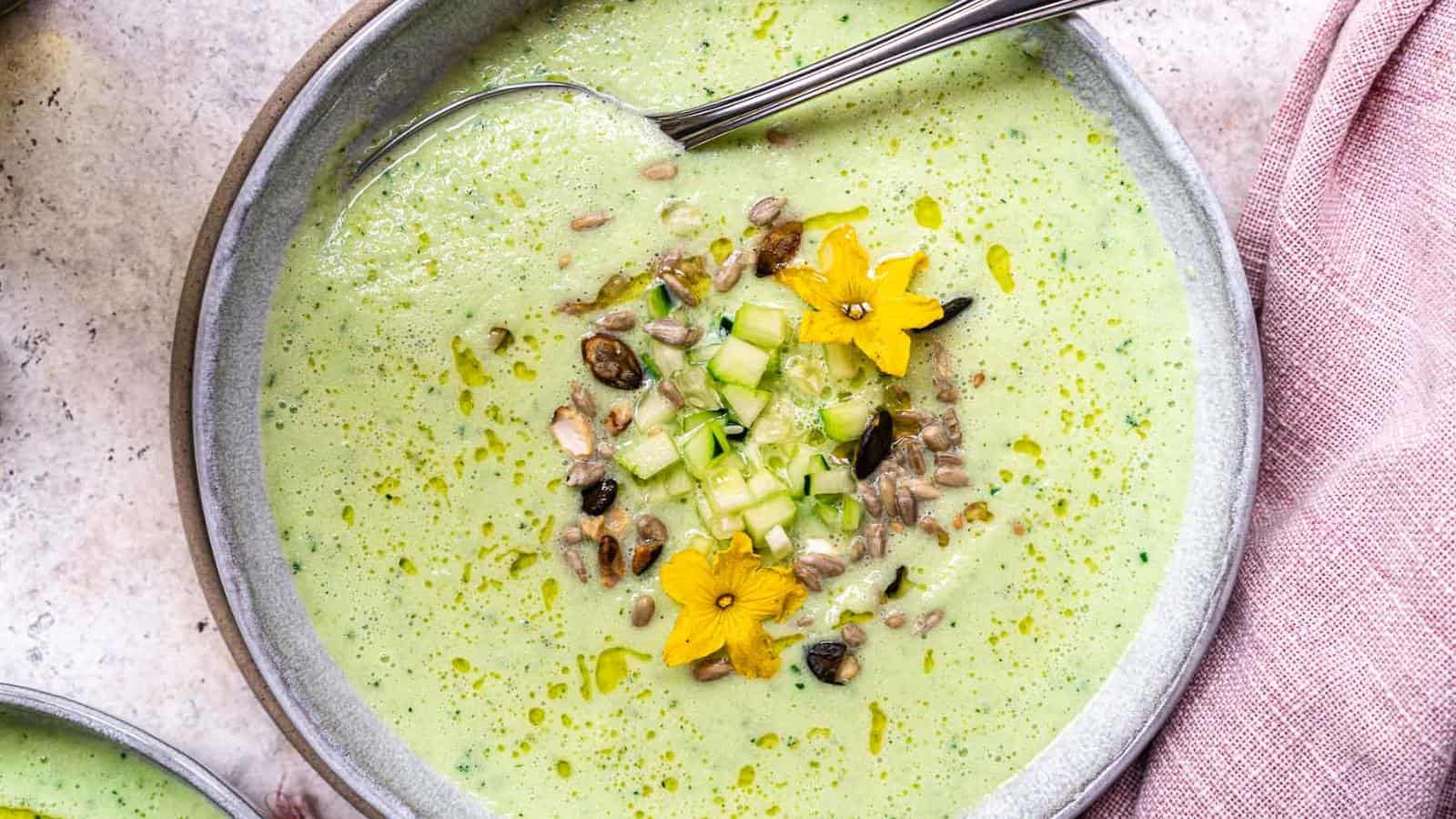 A bowl of green soup garnished with diced cucumber, seeds, yellow edible flowers, and a drizzle of oil, with a spoon resting in the bowl.