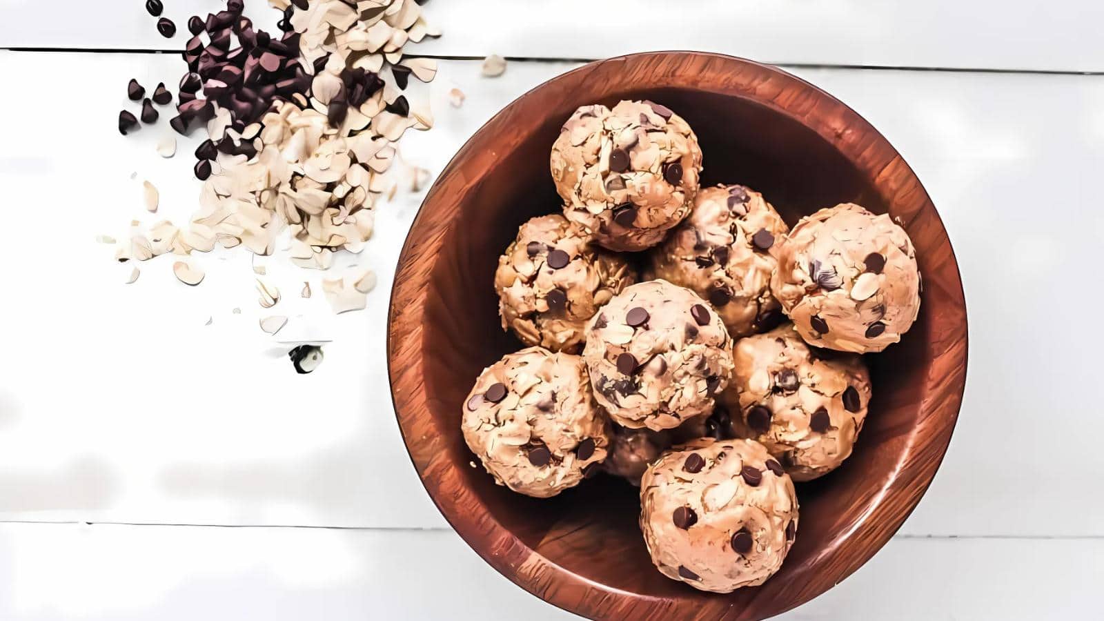 A wooden bowl filled with oatmeal and chocolate chip energy balls sits on a white surface with scattered chocolate chips and almond slices nearby.