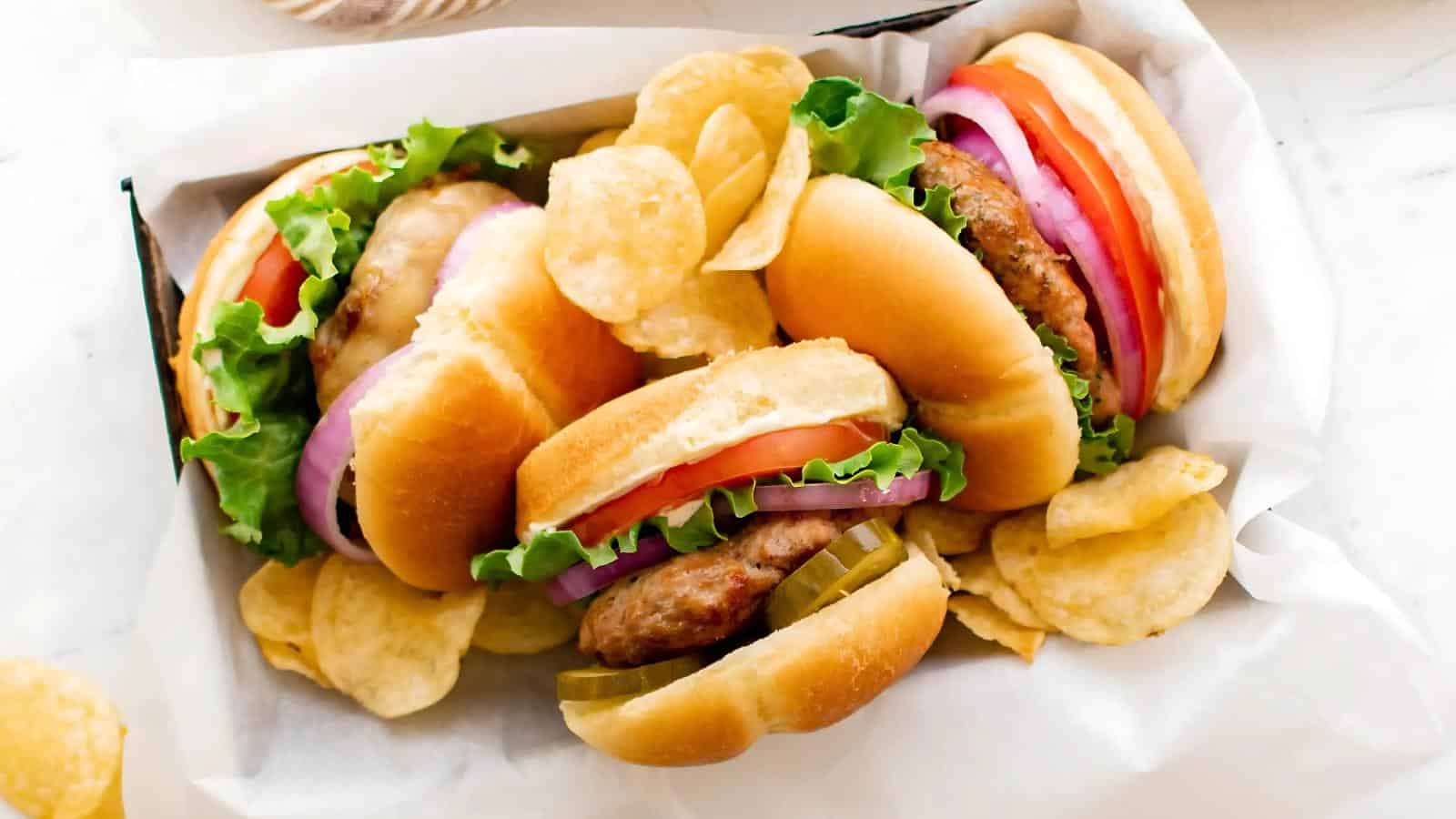 Three hamburgers with lettuce, tomato, onion, and pickles are served in a basket with potato chips on a white surface.