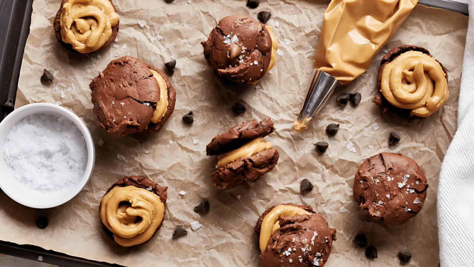 Chocolate cookies with a peanut butter filling are arranged on parchment paper with a piping bag, chocolate chips, and a bowl of salt scattered around.