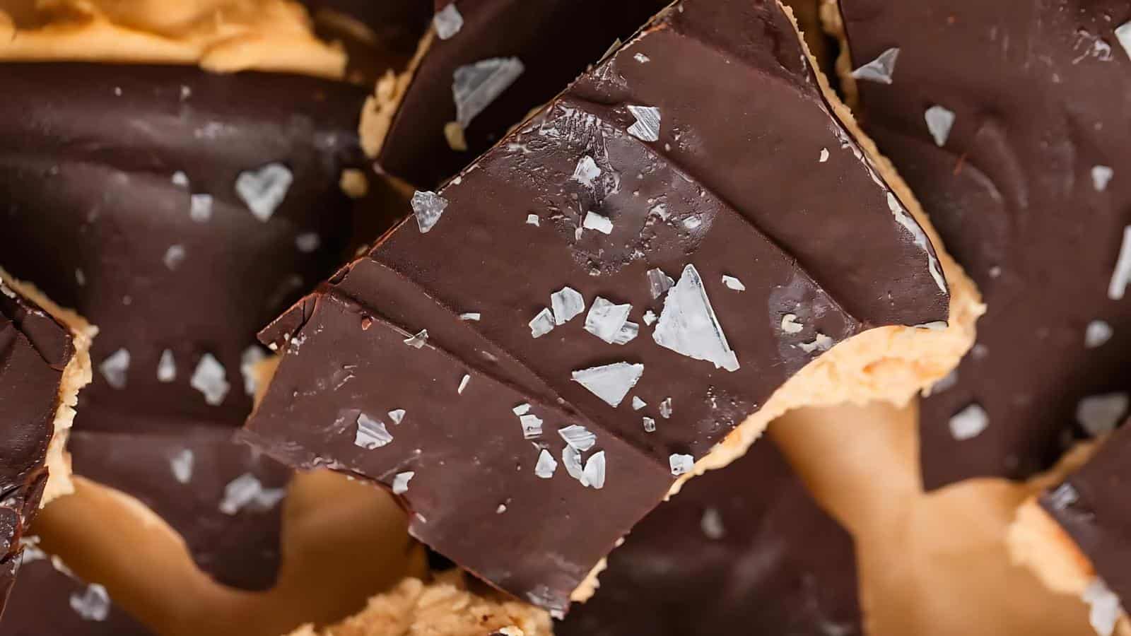 Close-up of several pieces of chocolate bark topped with flaky sea salt, revealing a creamy, tan-colored layer underneath the dark chocolate.