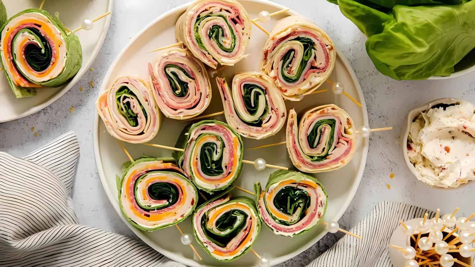 A plate of colorful pinwheel sandwiches with visible layers of deli meat, cheese, spinach, and tortillas, arranged neatly with toothpicks.