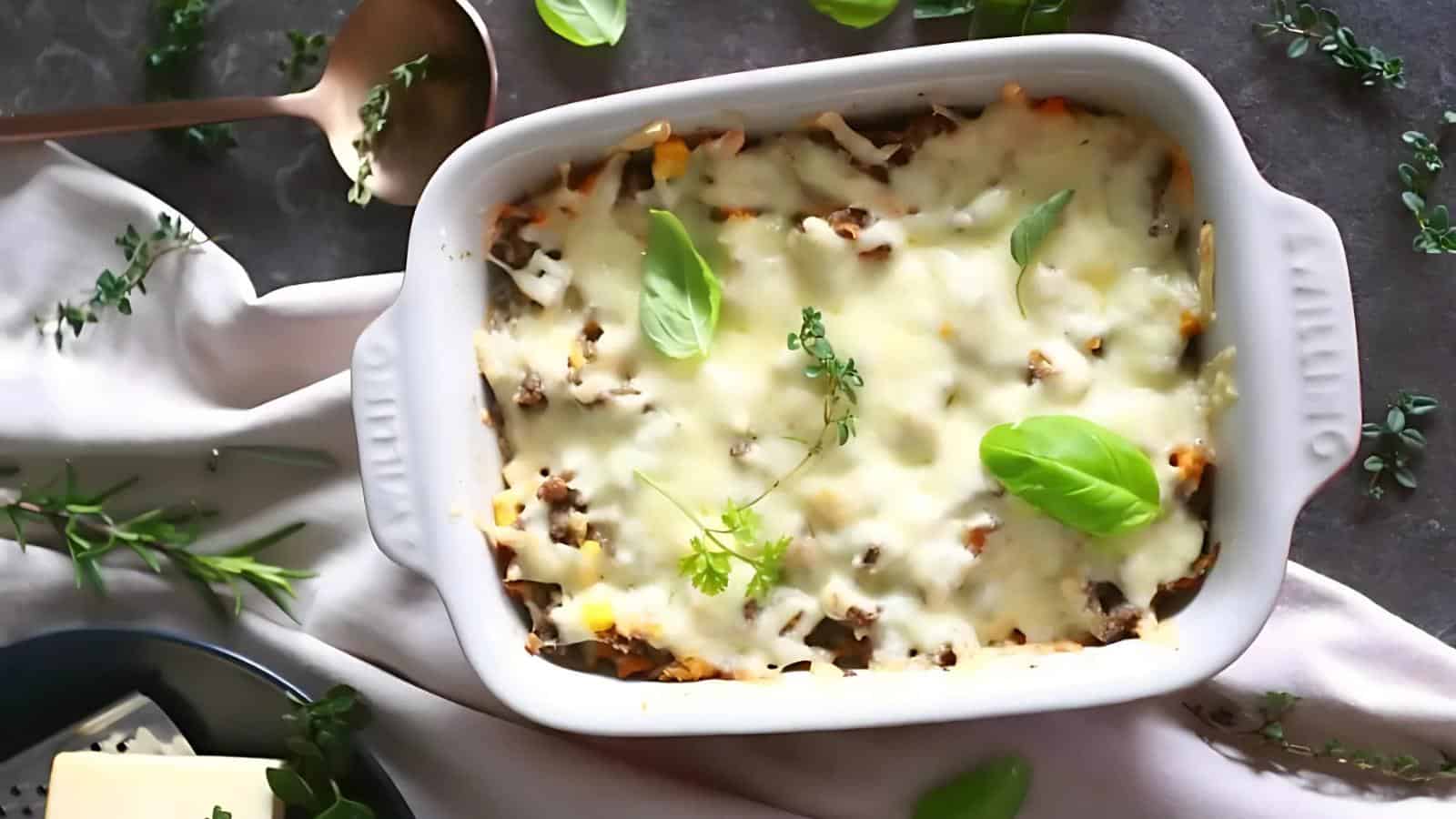 A baked casserole topped with melted cheese and garnished with fresh herbs sits in a white dish on a table with a spoon and scattered herb sprigs.