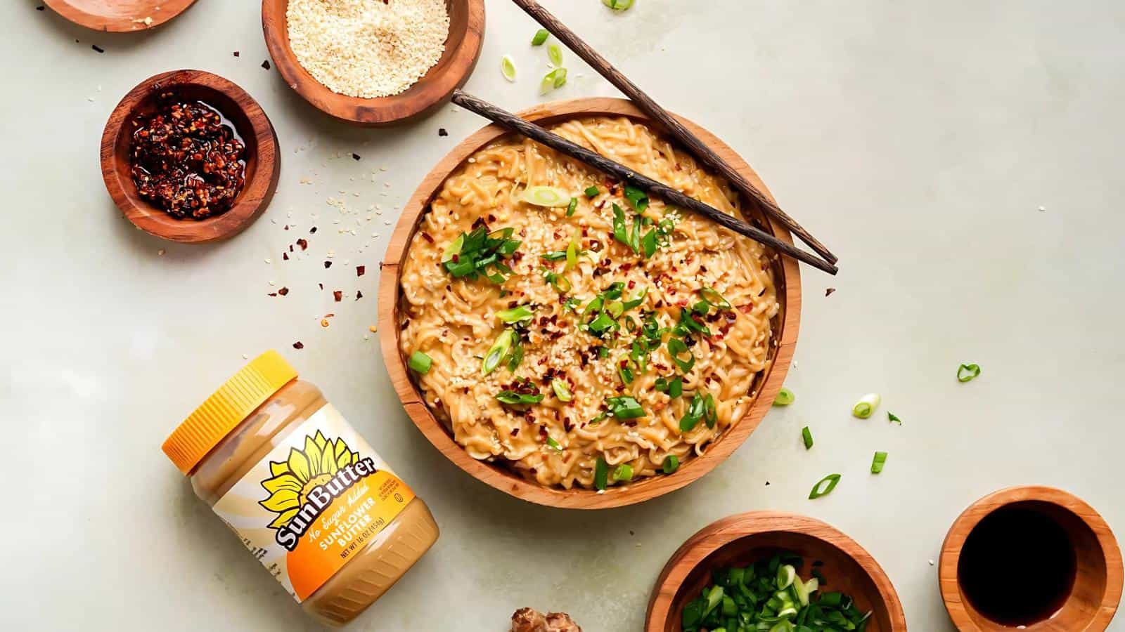 A bowl of noodles topped with chopped green onions and red pepper flakes, surrounded by seasonings, wooden bowls, chopsticks, and a jar of sunflower butter.