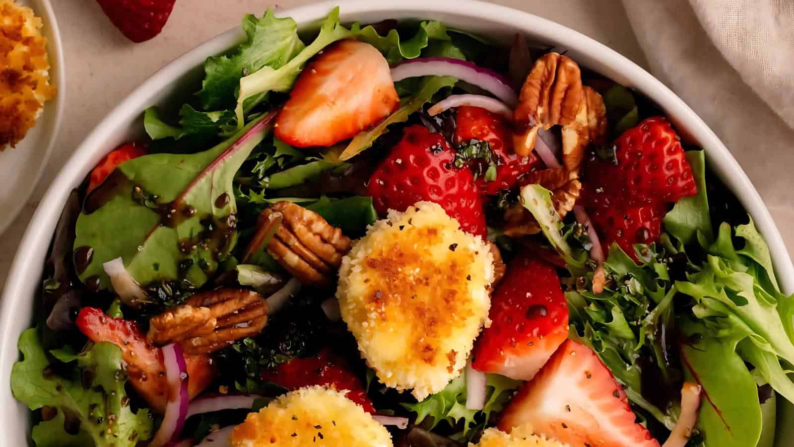 A bowl of mixed greens salad topped with sliced strawberries, pecans, red onion, and round pieces of breaded cheese.
