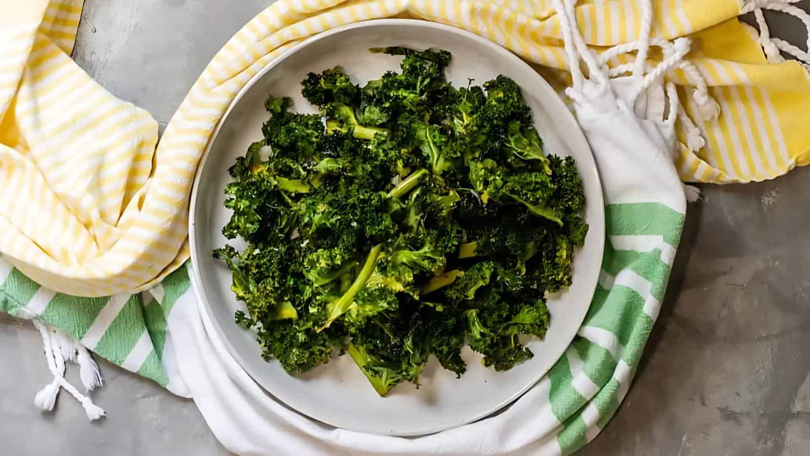 A white plate of cooked kale sits on a gray surface, surrounded by a yellow-striped towel and a green-striped towel.
