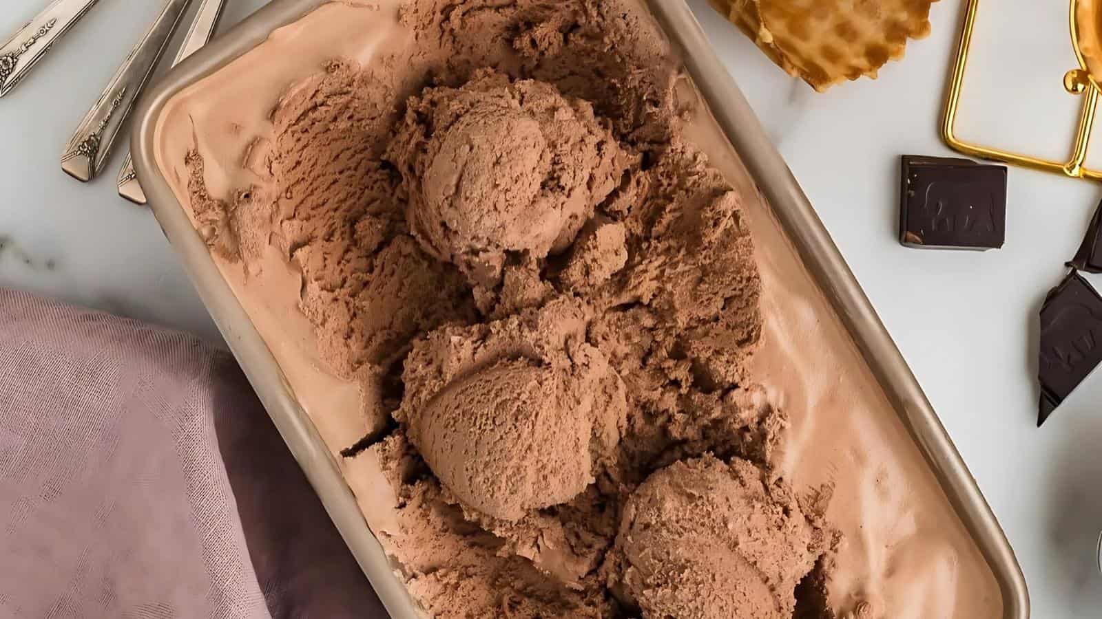 Rectangular container filled with chocolate ice cream, with scoops taken out. Pieces of waffle cone and chocolate are on the side, along with a spoon and napkin.