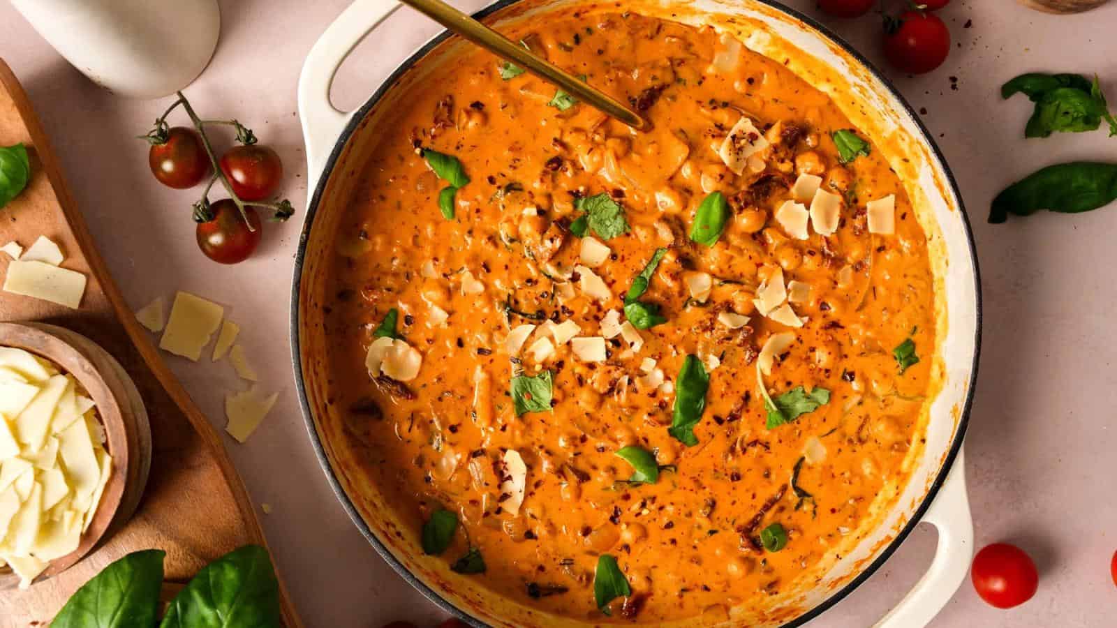 Large pot of creamy tomato basil pasta sauce garnished with fresh basil leaves and shaved cheese, surrounded by cherry tomatoes and cheese slices on a light surface.