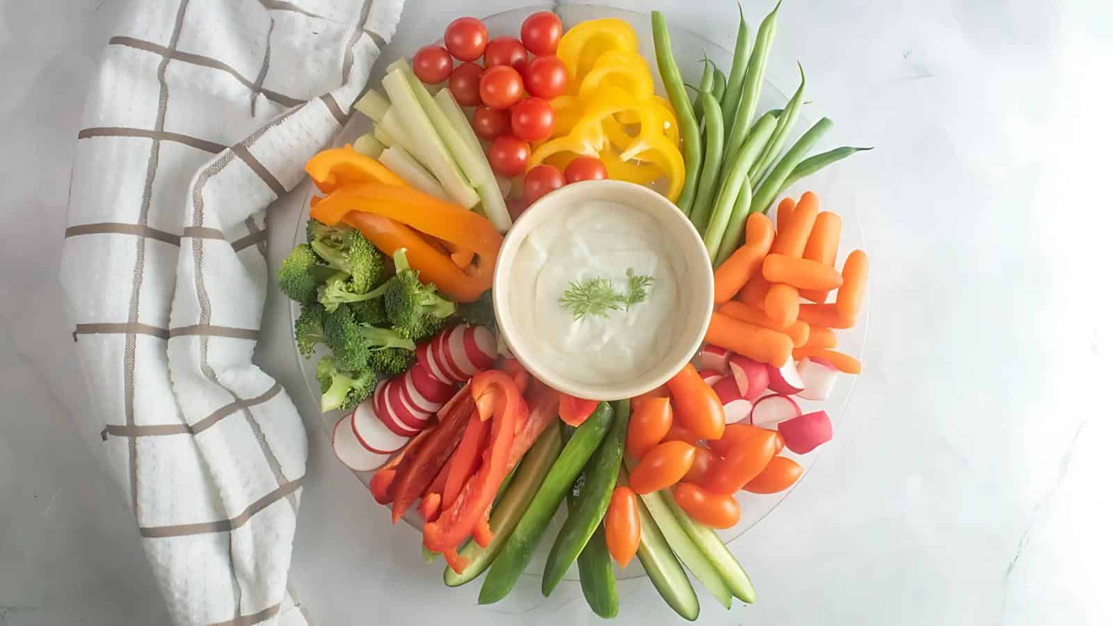 A platter of assorted fresh vegetables including carrots, bell peppers, cherry tomatoes, broccoli, cucumbers, and celery, arranged around a bowl of dip. A striped towel is nearby.