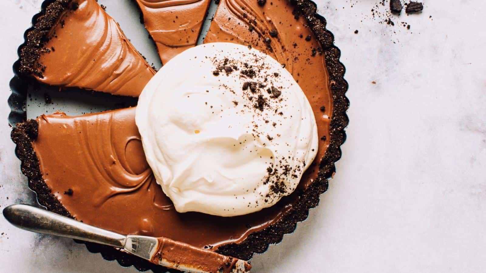 A chocolate tart with a cookie crust, topped with whipped cream, has several slices cut out and a knife resting on the edge.