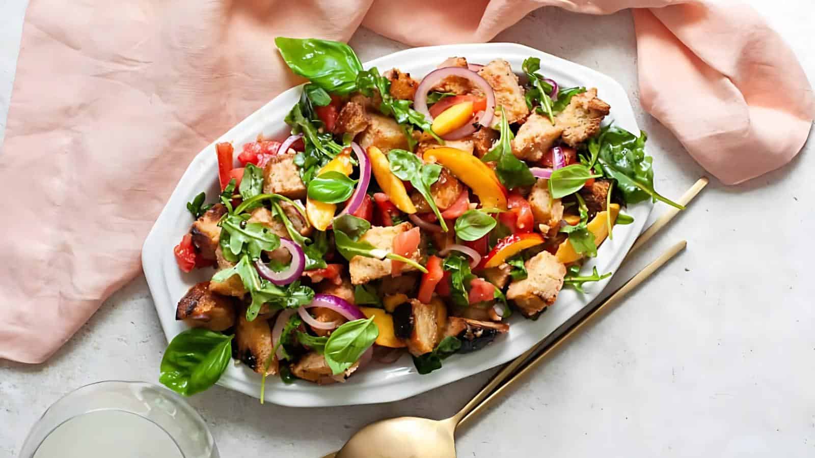 A white oval plate with panzanella salad, featuring chunks of bread, tomatoes, red onion, basil, arugula, and yellow bell pepper. Gold utensils and a pink napkin are nearby.