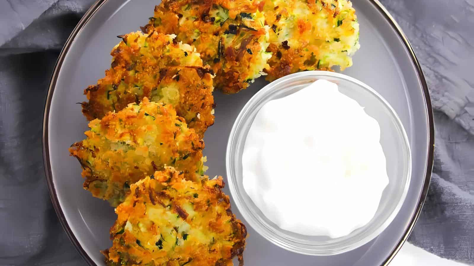 Four golden-brown vegetable fritters are arranged on a gray plate beside a small glass bowl of white dipping sauce.