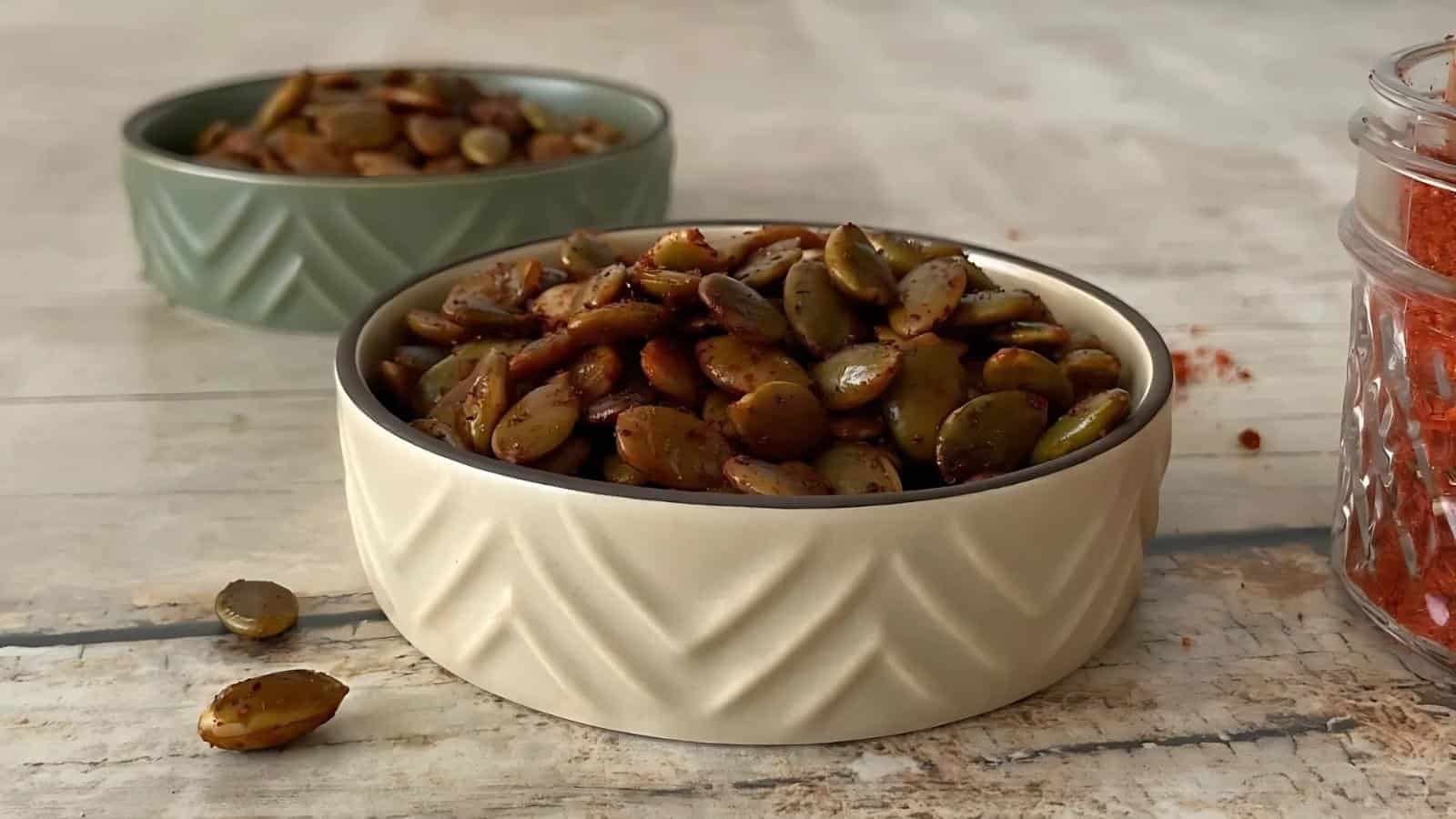 A bowl filled with roasted, seasoned pumpkin seeds is placed on a wooden surface, with another bowl and a jar of red spice in the background.