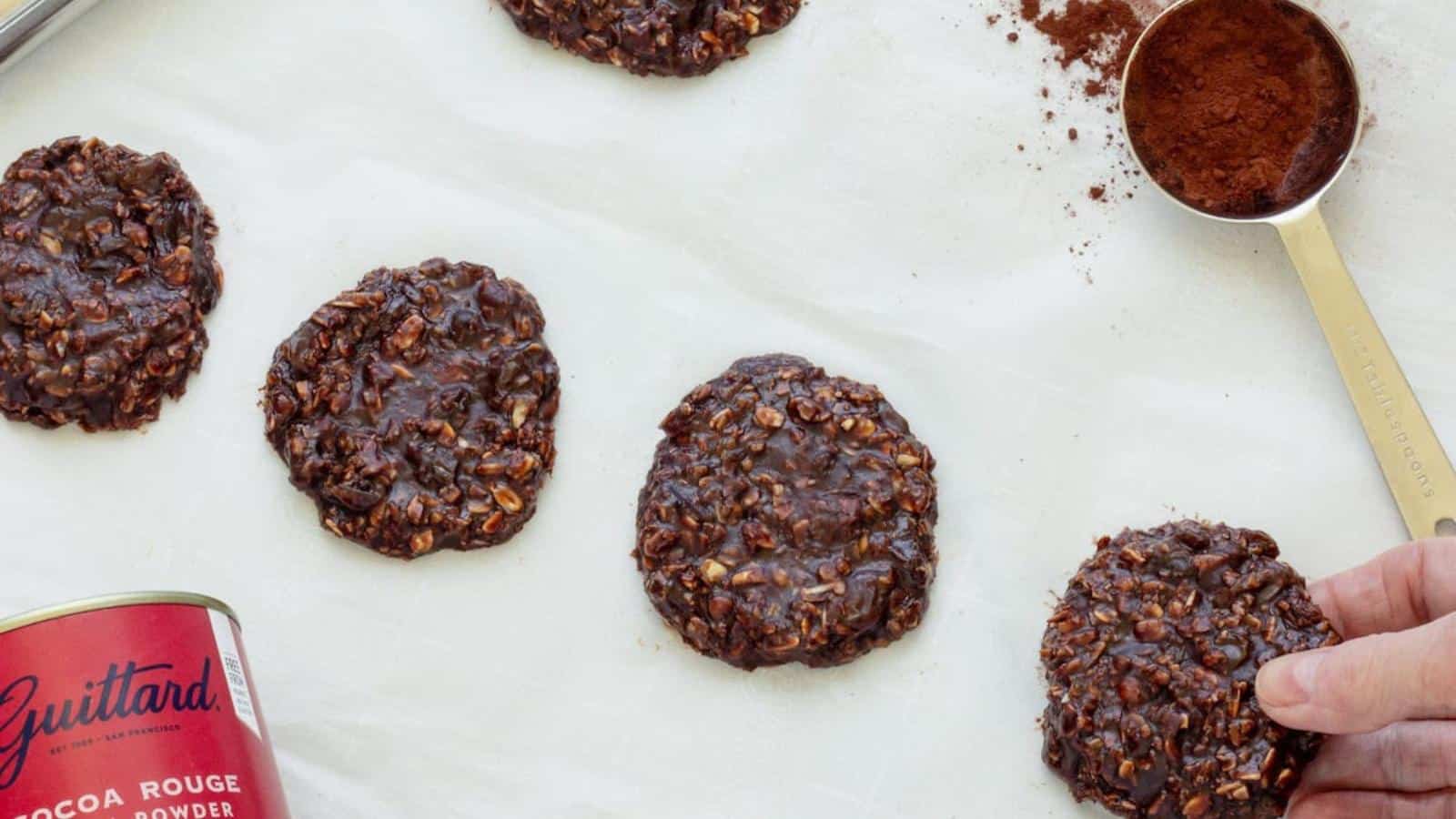 Several chocolate oatmeal cookies are arranged on a white surface next to a tin of cocoa powder, a measuring spoon with cocoa, and a hand reaching for one cookie.