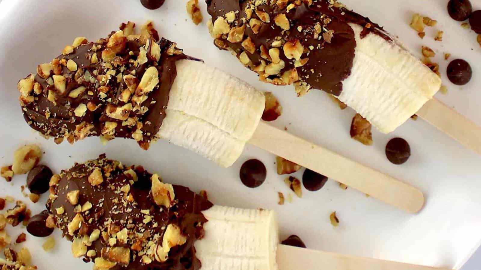 Three banana halves on sticks, partially dipped in chocolate and sprinkled with chopped nuts, are arranged on a white surface with chocolate chips scattered around.