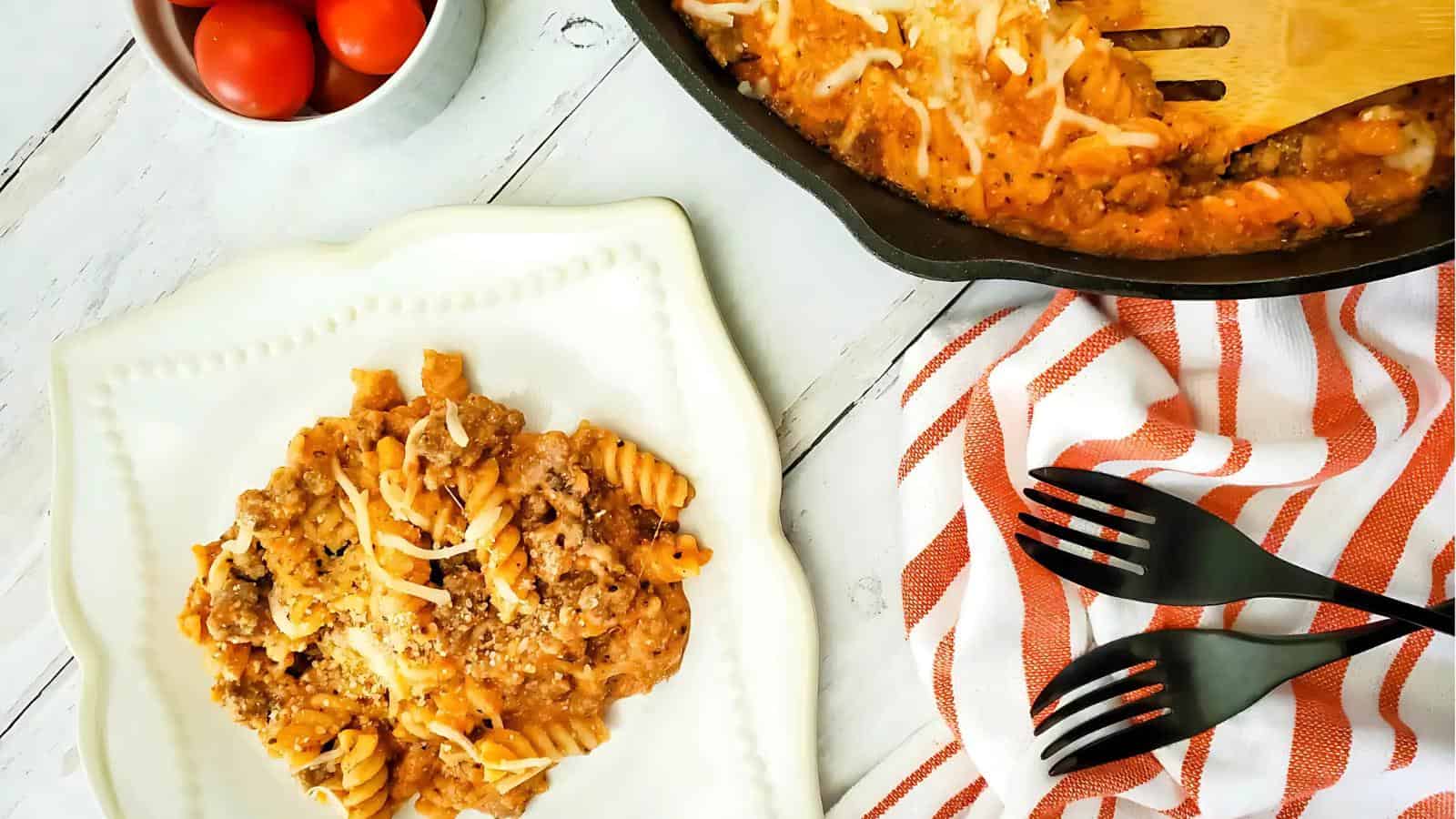 A plate of cheesy pasta with ground meat sits next to a skillet of the same dish, with tomatoes in a bowl and black forks on a striped towel nearby.