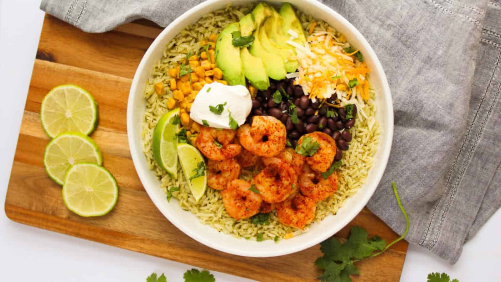 A bowl of rice topped with shrimp, avocado, black beans, corn, shredded cheese, and sour cream, garnished with lime wedges, on a wooden board with sliced limes and cilantro.