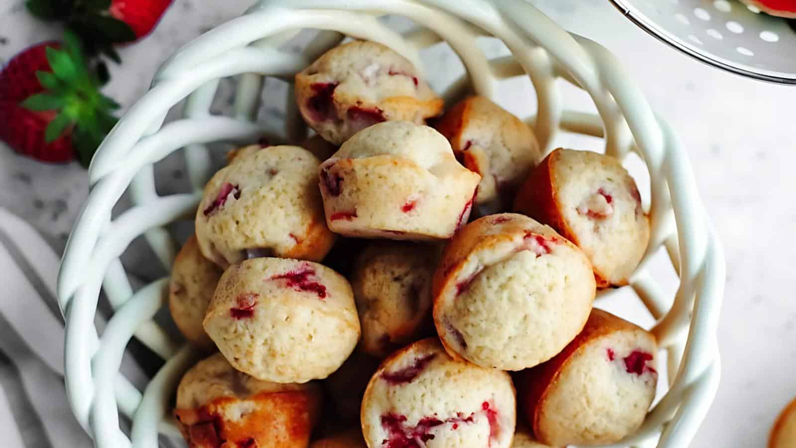 A white basket filled with mini muffins containing visible strawberry pieces, set on a light surface with fresh strawberries nearby.