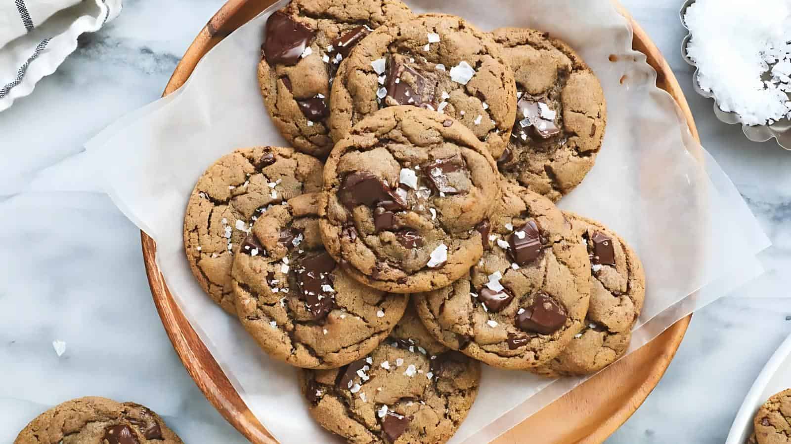 A wooden plate lined with parchment paper holds several chocolate chip cookies topped with flaky sea salt.