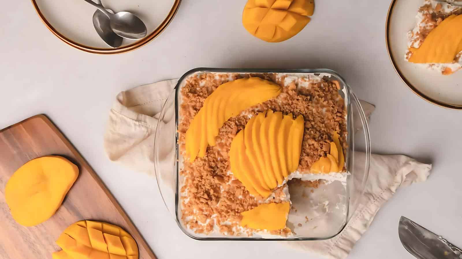 A glass dish of mango float dessert topped with sliced mango and crumbs, with plates, spoons, a cutting board, and mango slices arranged around it on a light surface.