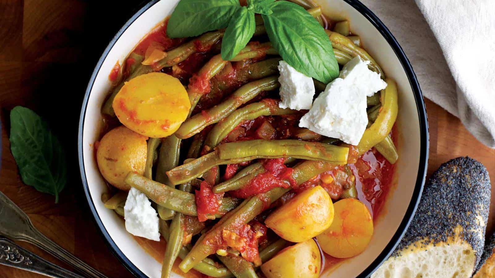 A bowl of green beans, potatoes, and tomatoes garnished with feta cheese and basil leaves, served with a slice of bread on the side.