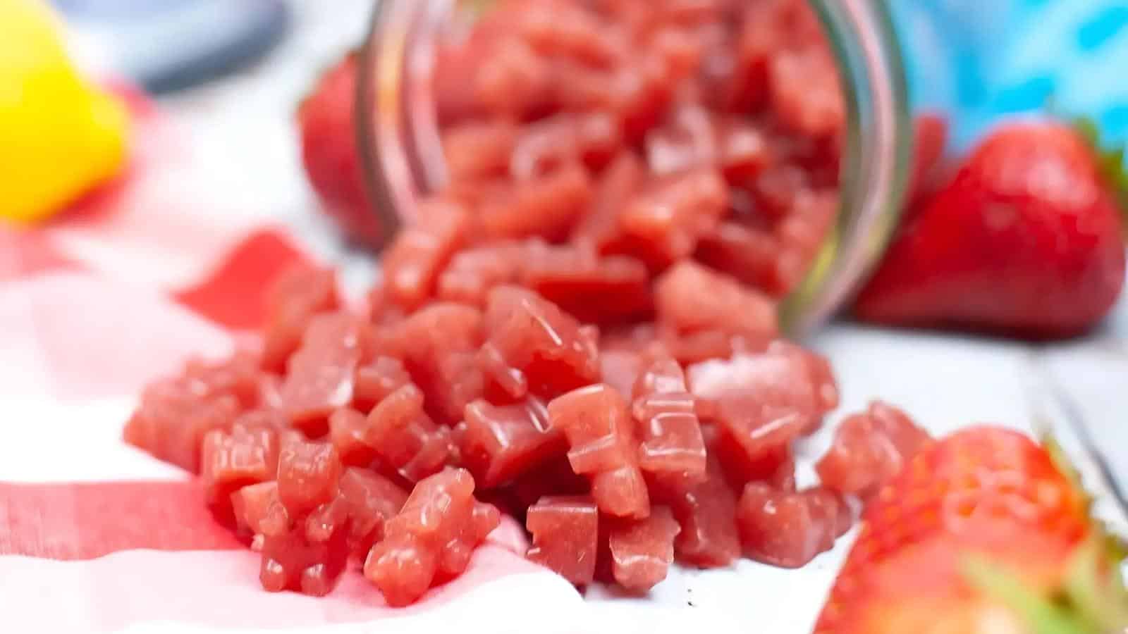 Close-up of small diced red fruit pieces spilling from a jar onto a striped surface, with whole strawberries and other blurred objects in the background.