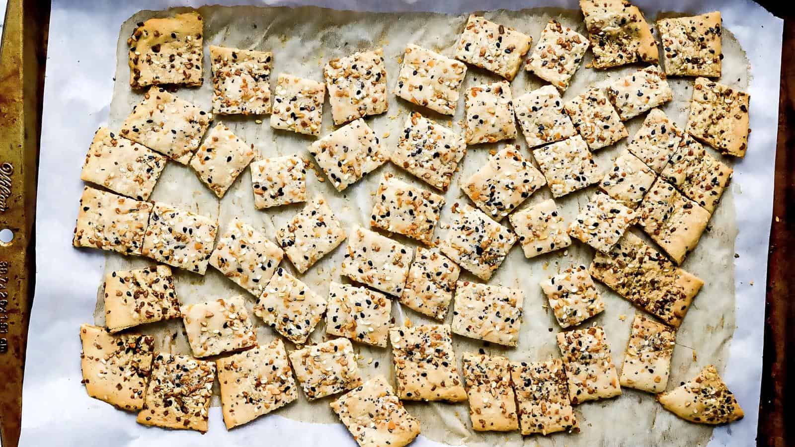 A baking sheet lined with parchment paper holds numerous rectangular, seed-topped crackers arranged in a scattered pattern.