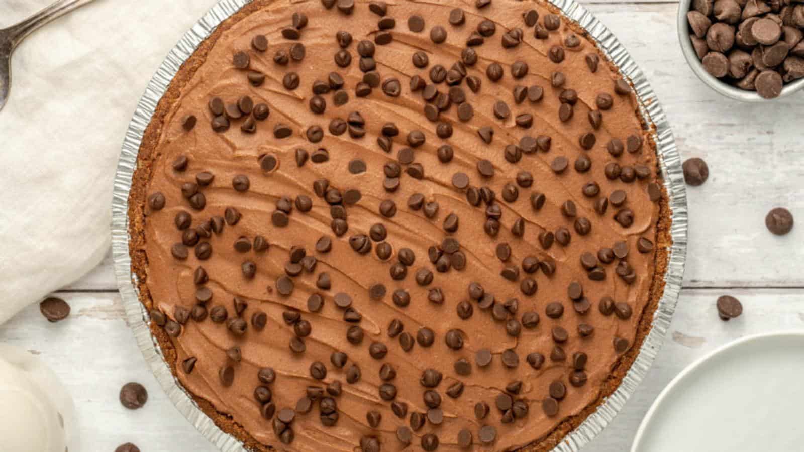 A chocolate cream pie in a foil pan topped with chocolate chips, shown on a white wooden surface with plates, a spoon, and a bowl of chocolate chips nearby.