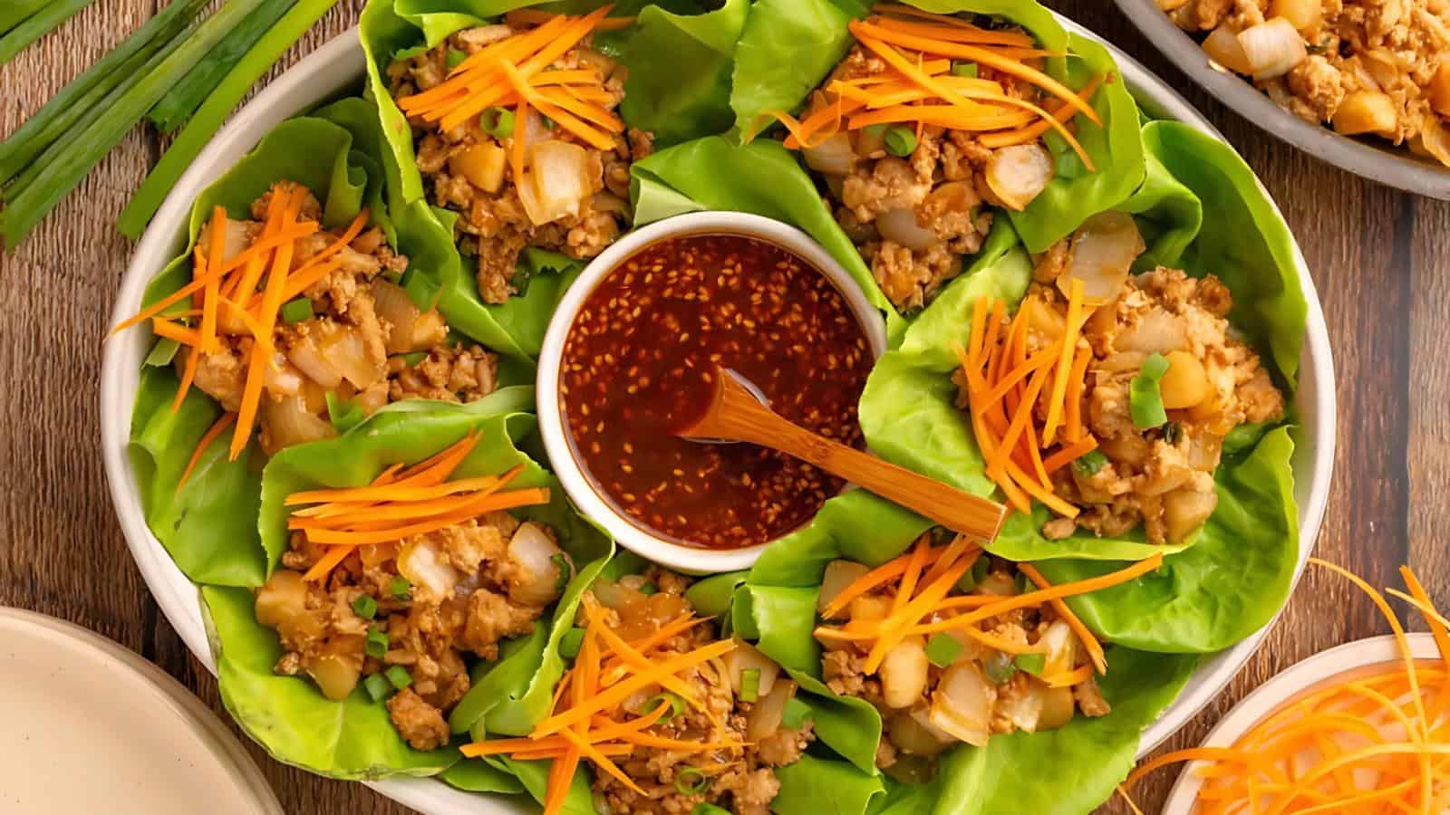 A plate of lettuce wraps filled with chopped vegetables and minced meat, topped with shredded carrots, arranged around a bowl of dipping sauce.